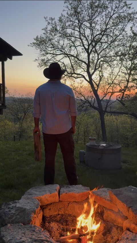 Standing at a lit fire pit as the sun sets over the hills